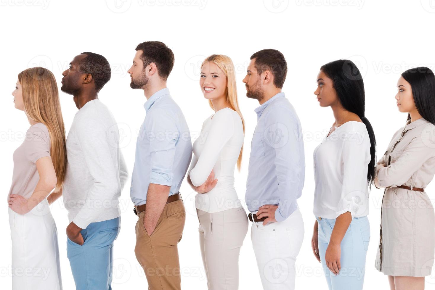 attendant son tour. vue latérale d'une belle jeune femme regardant la caméra et souriant tout en se tenant dans une rangée avec d'autres personnes et sur fond blanc photo