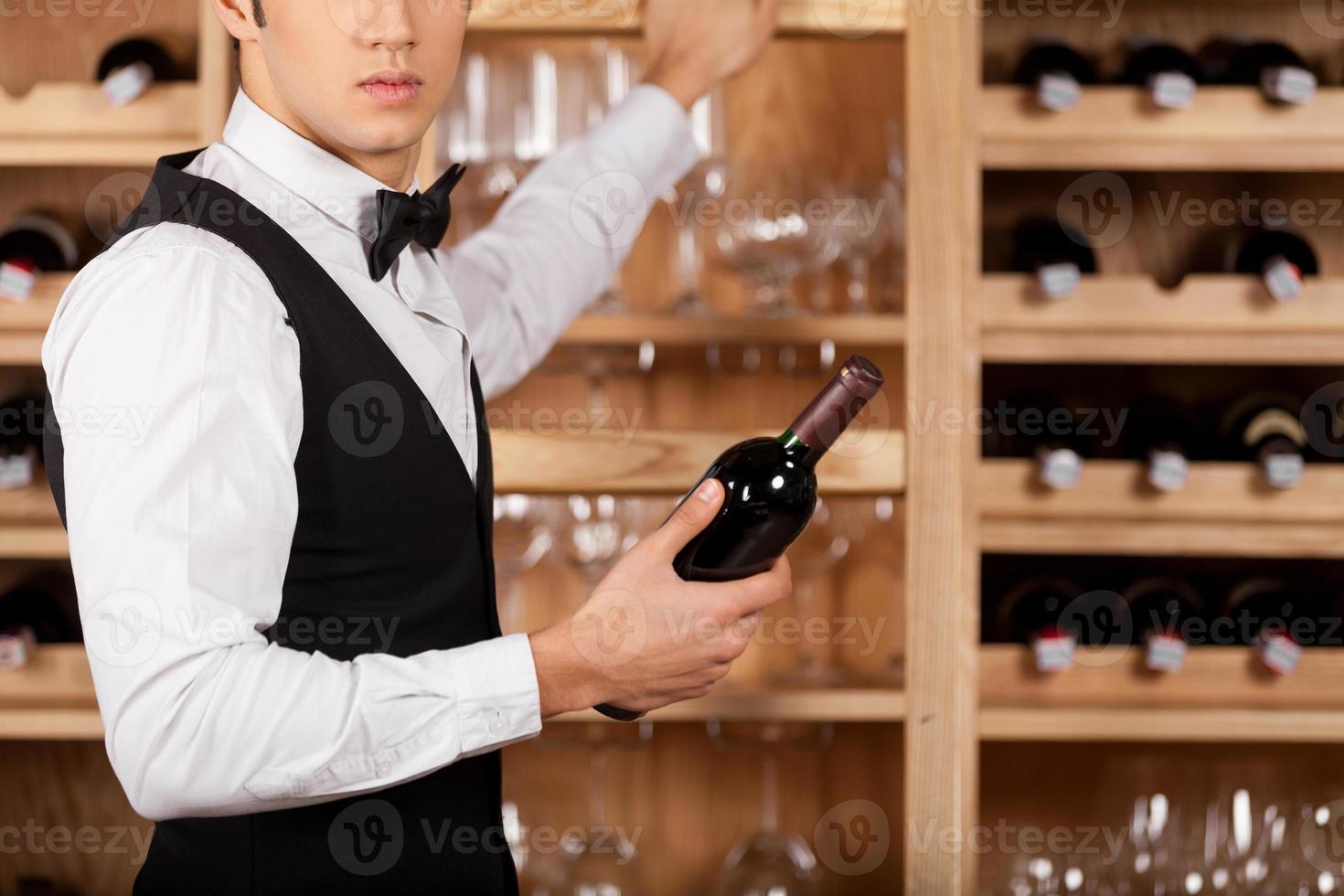 présentant le meilleur vin. image recadrée d'un jeune sommelier joyeux debout devant une étagère à vin et tenant une bouteille de vin photo