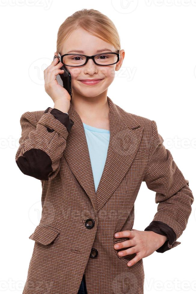 parler d'affaires. joyeuse petite fille en tenues de soirée parlant au téléphone mobile et tenant la main sur la hanche en se tenant debout isolé sur blanc photo