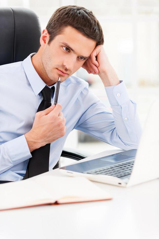 en attente d'inspiration. jeune homme réfléchi en chemise et cravate regardant un ordinateur portable et touchant son menton avec un stylo alors qu'il était assis sur son lieu de travail photo