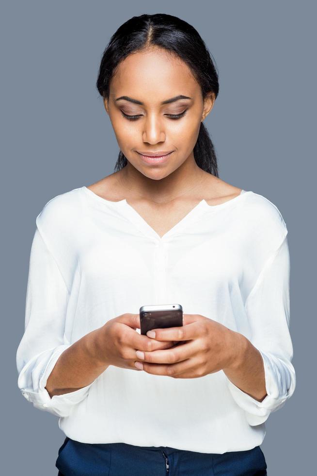 taper un message à un ami. confiante jeune femme africaine tenant un téléphone portable et le regardant avec le sourire en se tenant debout sur fond gris photo