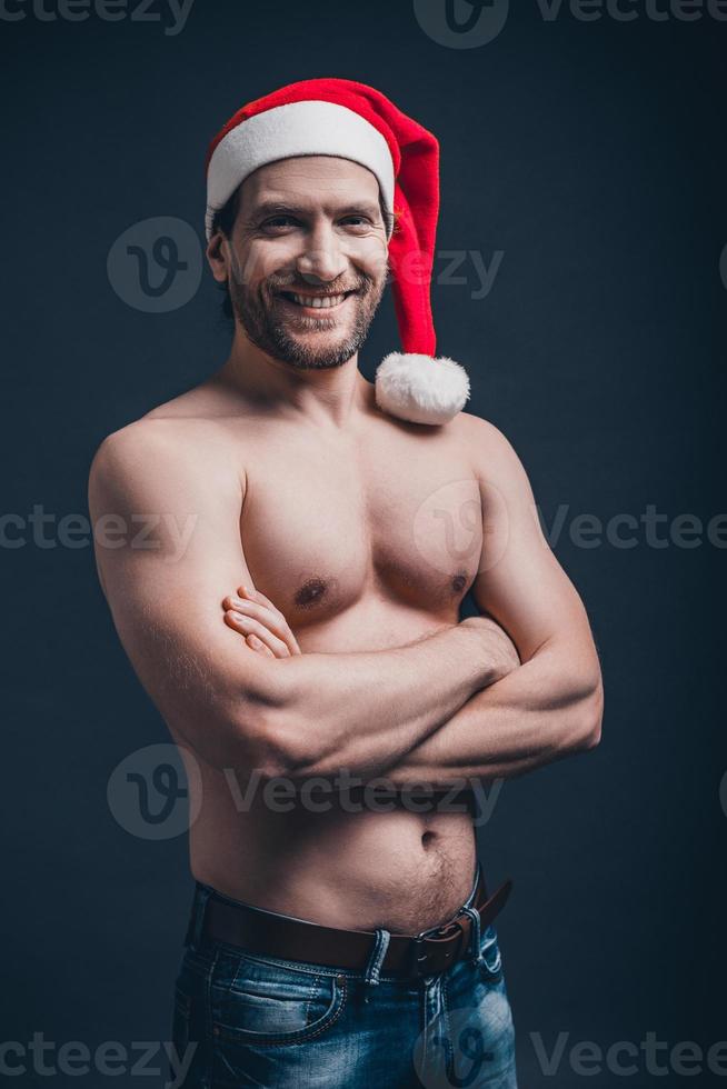 père noël sexy. jeune homme confiant, gardant les bras croisés et regardant la caméra en se tenant debout sur fond noir photo