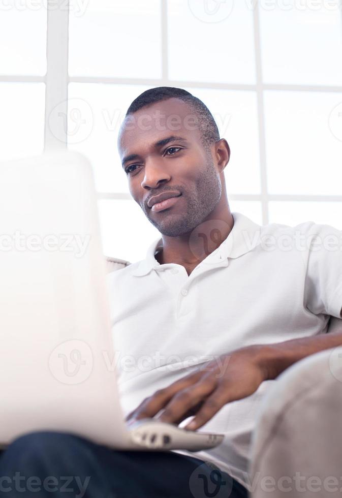 travailler à la maison. beau jeune homme africain utilisant un ordinateur et assis sur la chaise photo