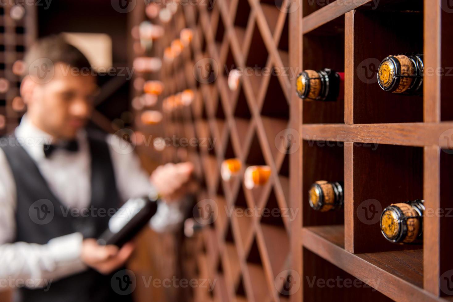 je recommande ce vin. sommelier masculin confiant montrant une bouteille de vin et souriant tout en se tenant près de l'étagère à vin photo