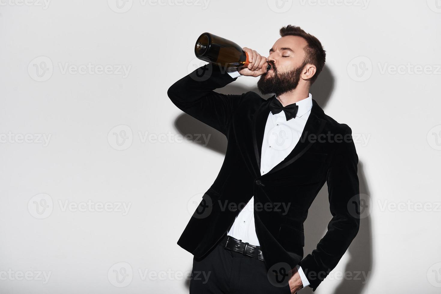 célébrer quelque chose de spécial. beau jeune homme en costume complet buvant du champagne de la bouteille en se tenant debout sur fond gris photo