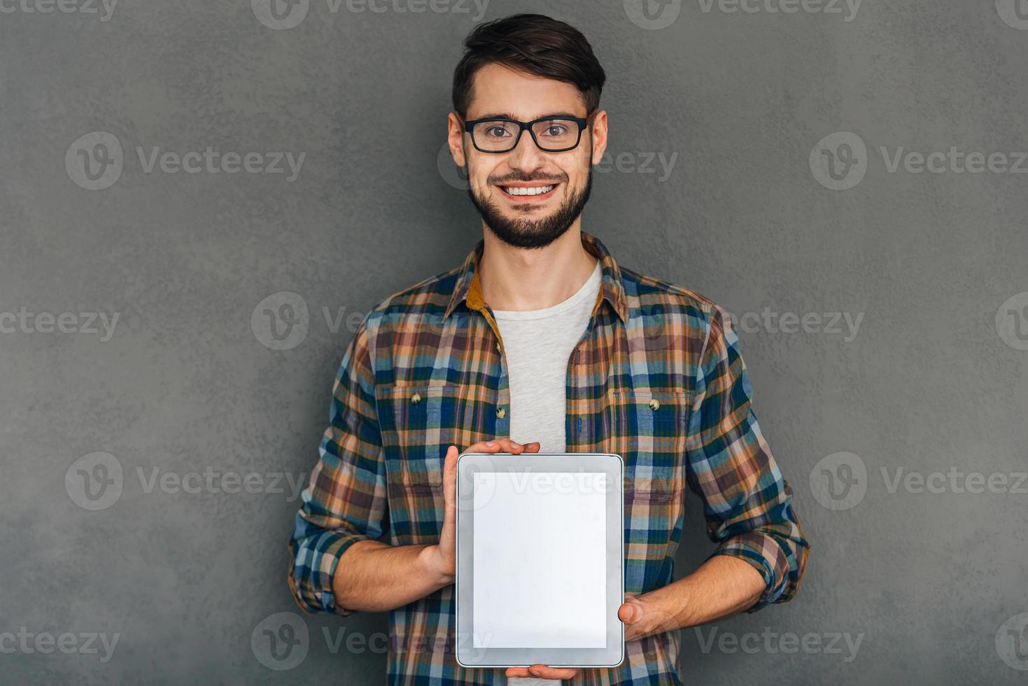 cela peut vous aider un jeune homme joyeux à lunettes montrant sa tablette numérique avec espace de copie et regardant la caméra avec le sourire en se tenant debout sur fond gris photo