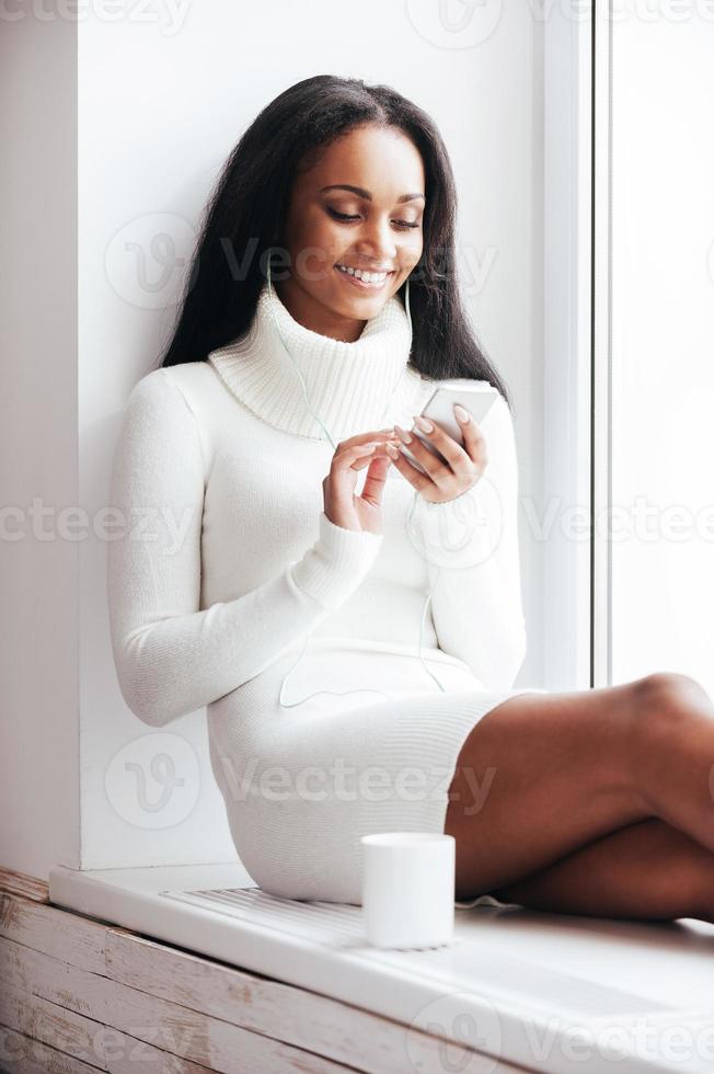 profiter de la musique et du café chaud. belle jeune femme africaine au casque écoutant de la musique et regardant son lecteur mp3 assis sur le rebord de la fenêtre photo