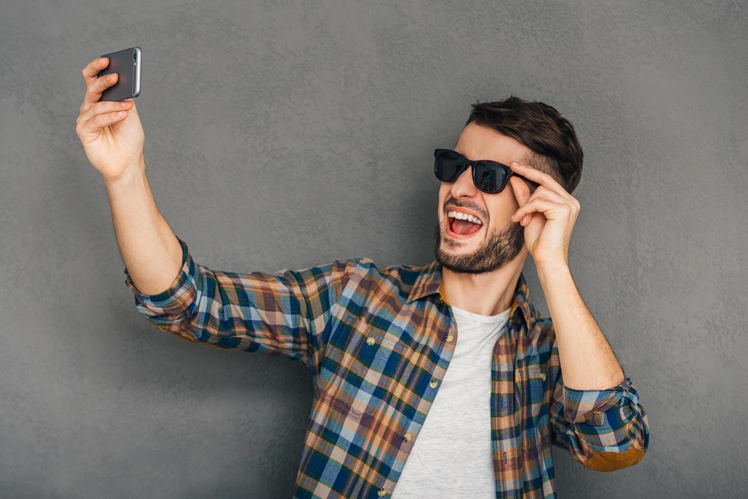 le temps des selfies. joyeux jeune homme à lunettes de soleil faisant selfie en se tenant debout sur fond gris photo