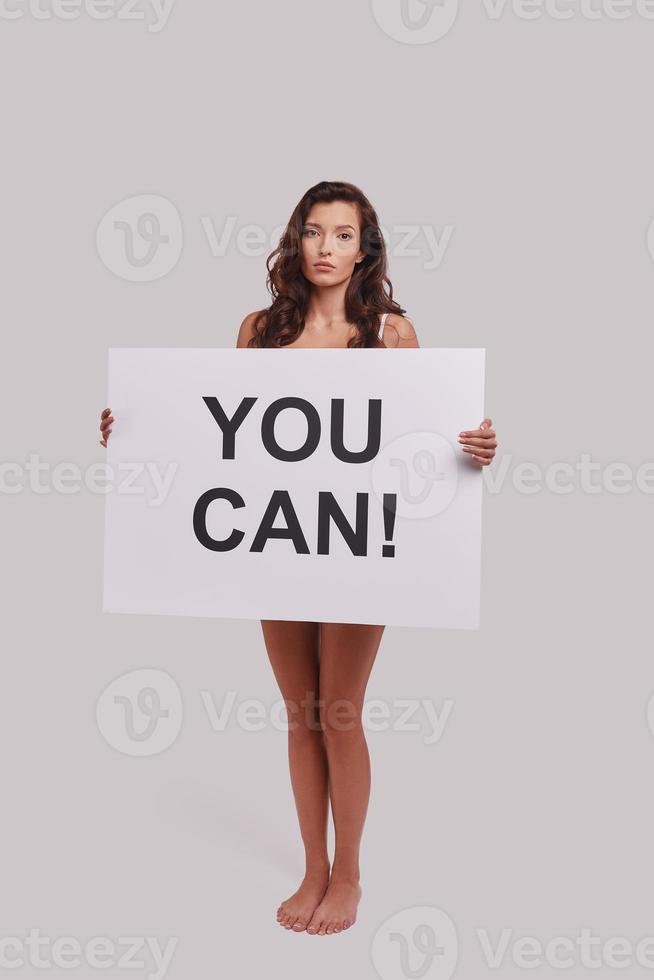 toute la longueur de la jolie jeune femme couvrant avec une affiche et regardant la caméra en se tenant debout sur fond gris photo