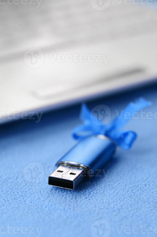 carte mémoire flash usb bleu brillant avec un arc bleu se trouve sur une couverture de tissu polaire bleu clair doux et poilu à côté d'un ordinateur portable blanc. conception de cadeau féminin classique pour une carte mémoire photo