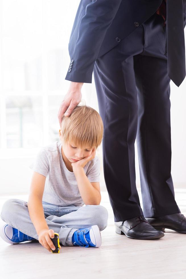 manque son père occupé. triste petit garçon tenant la main sur le menton tout en jouant avec une petite voiture pendant que son père en tenue de soirée le consolait photo