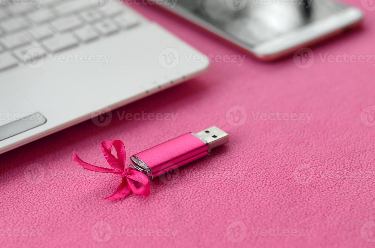 une carte mémoire flash usb rose brillante avec un arc rose se trouve sur une couverture en tissu polaire rose clair doux et poilu à côté d'un ordinateur portable et d'un smartphone blancs. conception de cadeau féminin classique pour une carte mémoire photo