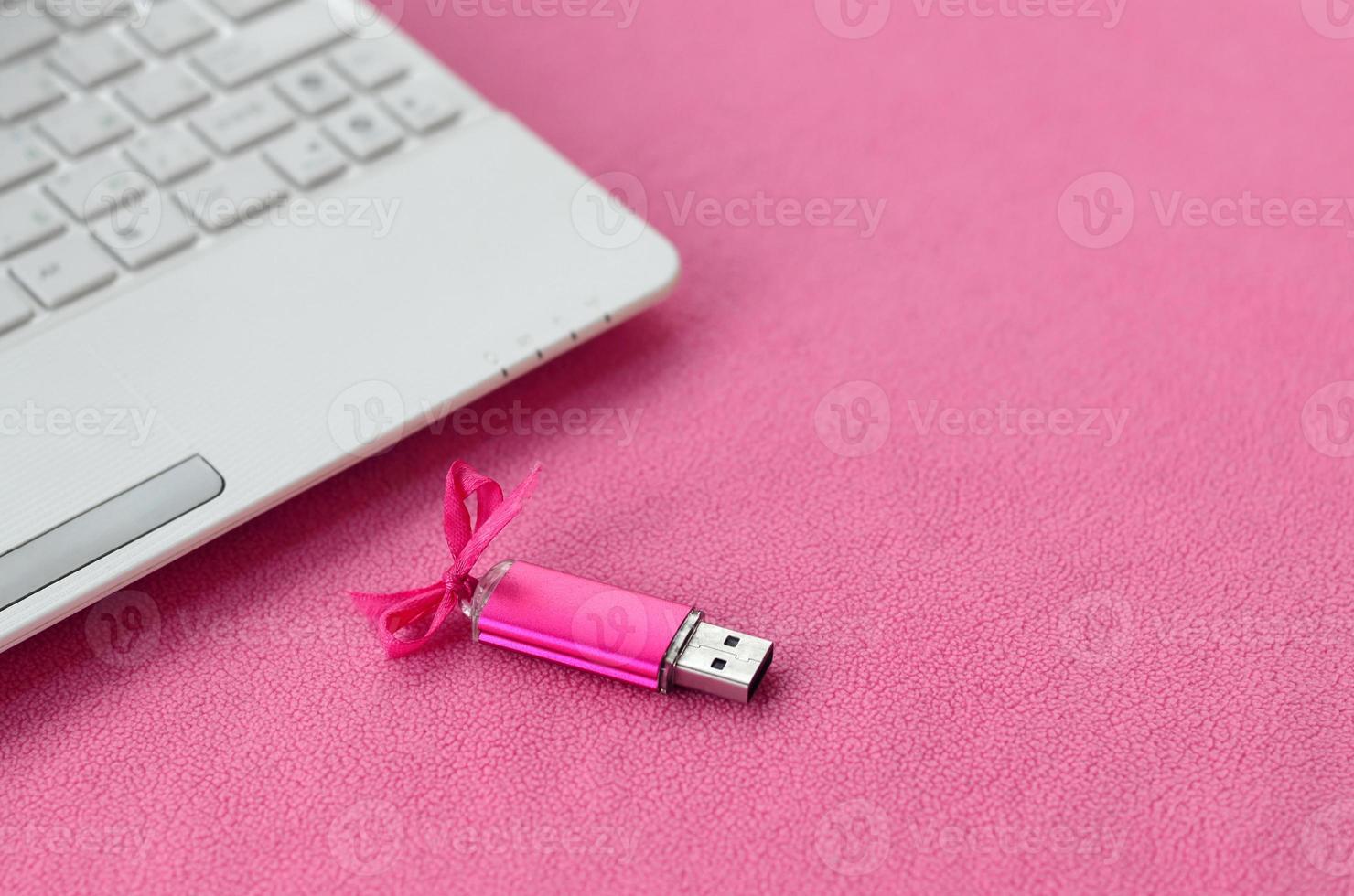 carte mémoire flash usb rose brillant avec un arc rose se trouve sur une couverture de tissu polaire rose clair doux et poilu à côté d'un ordinateur portable blanc. conception de cadeau féminin classique pour une carte mémoire photo