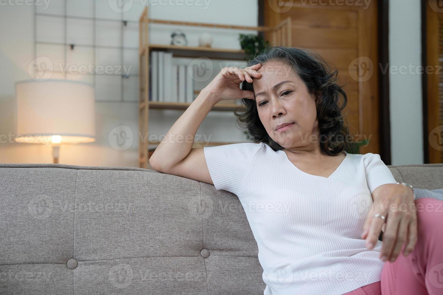 une femme senior asiatique déprimée s'assoit sur un canapé en regardant loin par la fenêtre. solitude femme âgée vivant seule à la maison avec un visage triste. concept de soins de santé mentale à la retraite et de rétablissement de la maladie photo