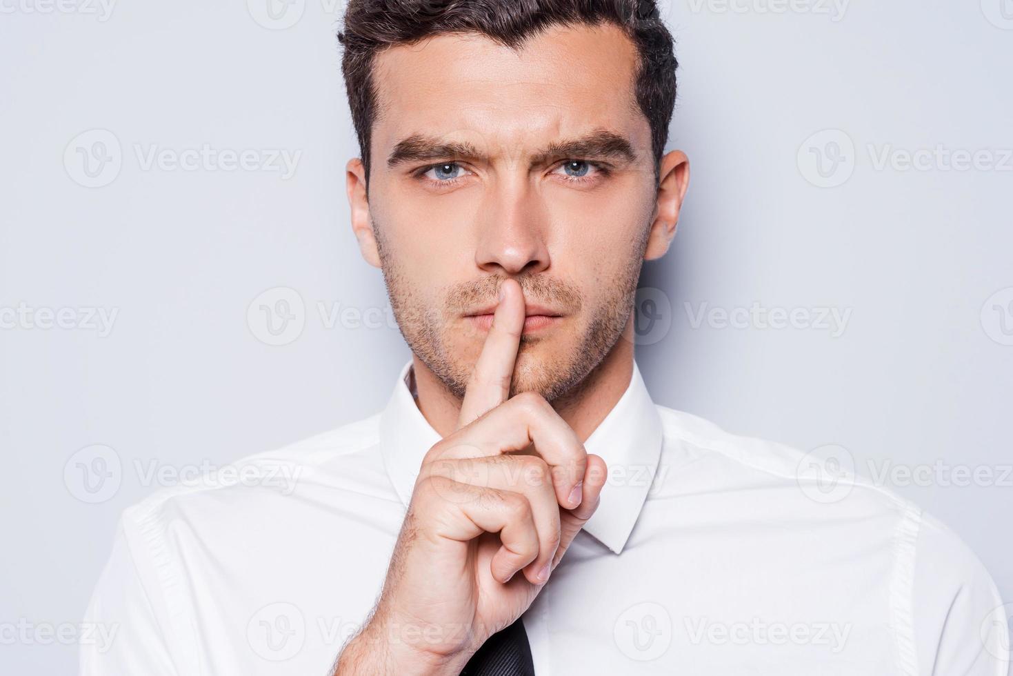 garder le silence confiant jeune homme en chemise blanche regardant la caméra et tenant le doigt sur les lèvres en se tenant debout sur fond gris photo