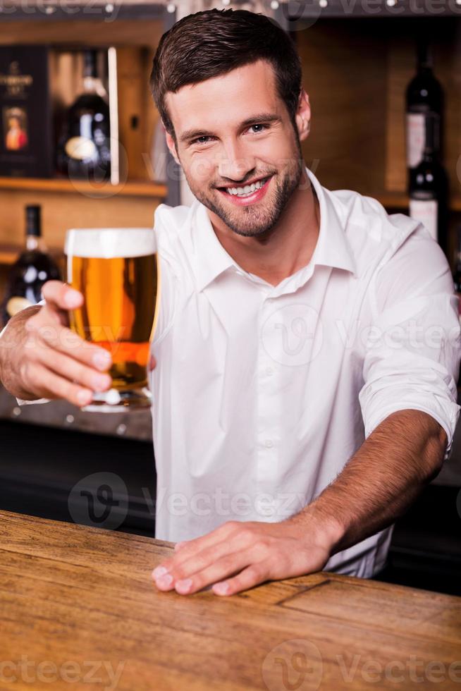 laissez-moi étancher votre soif beau jeune barman en chemise blanche tendant un verre avec de la bière et souriant tout en se tenant au comptoir du bar photo