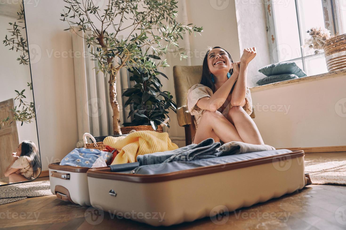 belle jeune femme à l'air heureuse assise près de la valise ouverte sur le sol à la maison photo