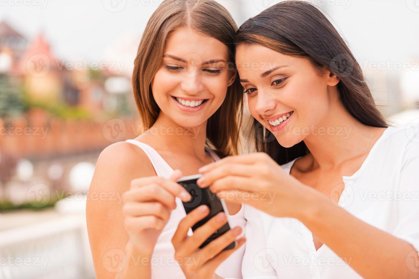 regardez ces deux belles jeunes femmes regardant un téléphone portable et souriant tout en se tenant à l'extérieur ensemble photo