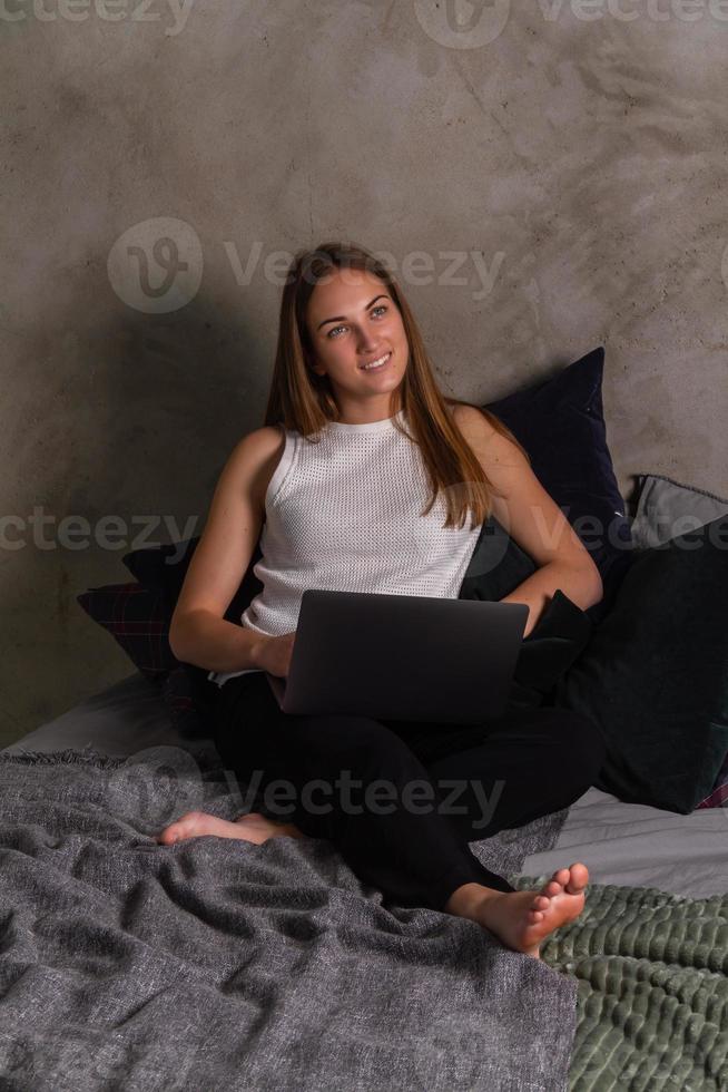 femme rêveuse regardant vers le haut assise sur le lit avec un ordinateur portable sur ses genoux photo