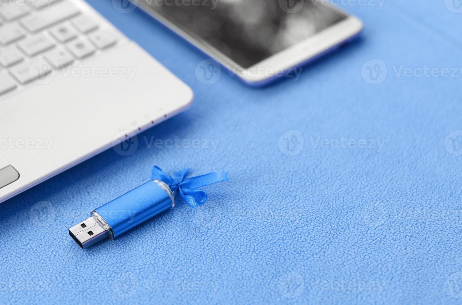 carte mémoire flash usb bleu brillant avec un arc bleu se trouve sur une couverture de tissu polaire bleu clair doux et poilu à côté d'un ordinateur portable blanc et d'un smartphone. conception de cadeau féminin classique pour une carte mémoire photo