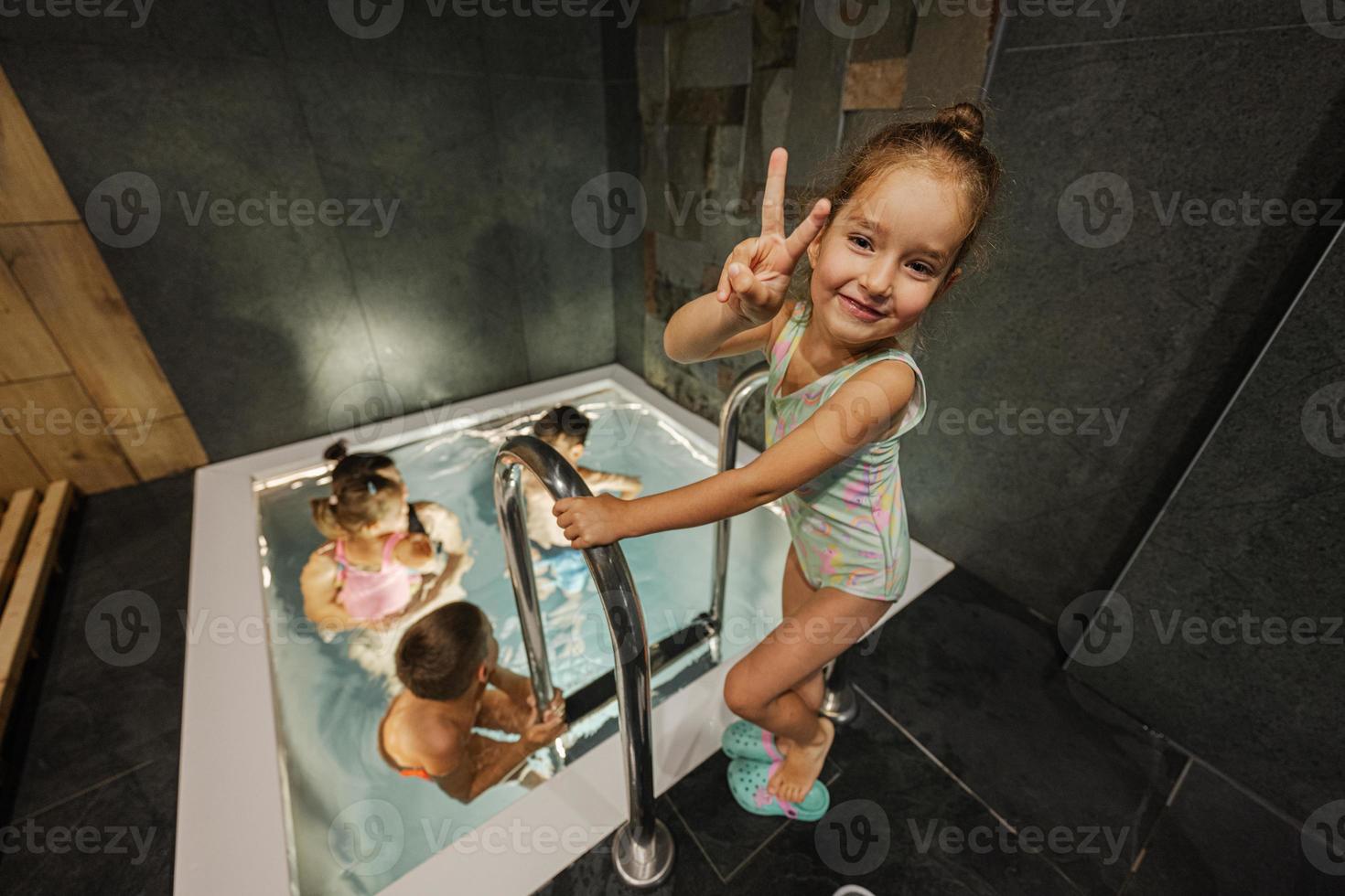 bébé fille montre deux doigts contre sa mère avec des enfants nageant dans la piscine du spa au sauna. photo