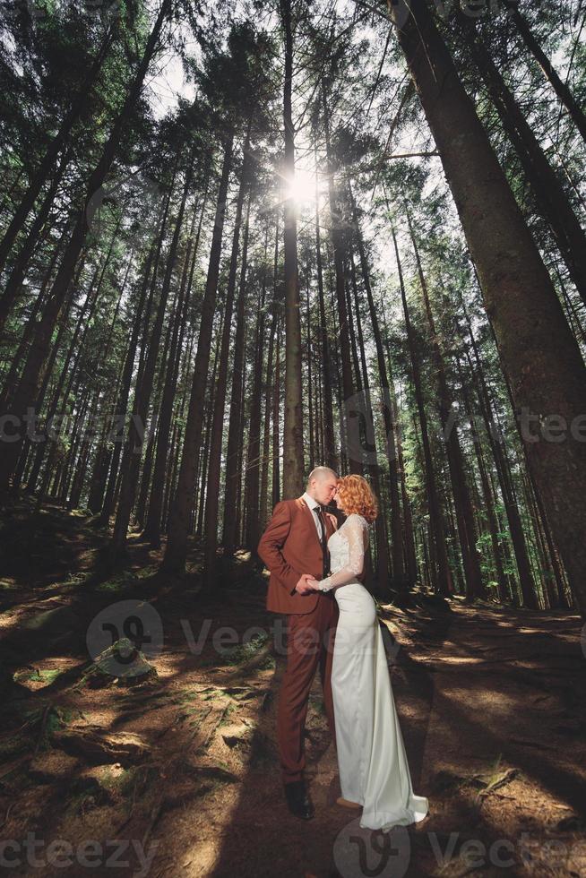 heureux couple élégant jeunes mariés dans la forêt verte le jour de l'été. la mariée en longue robe blanche et le marié en costume rouge s'étreignent. Jour de mariage. photo