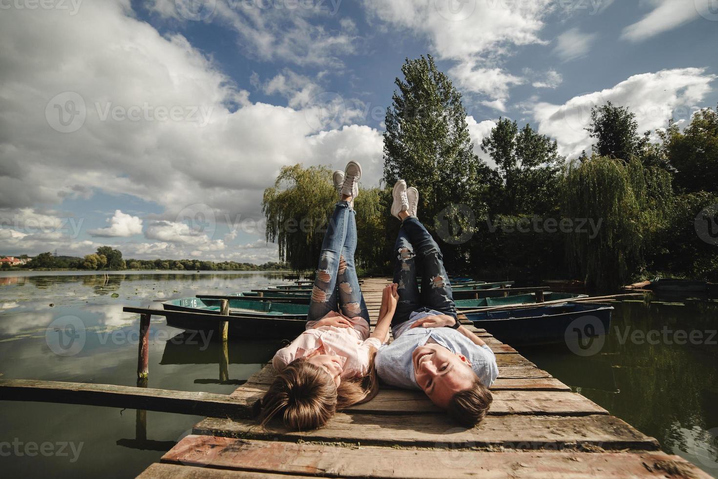 rencard parfait. beau jeune couple et souriant en position couchée sur la jetée. heureux jeune homme et femme riant en position couchée sur une jetée en bois près de l'eau photo