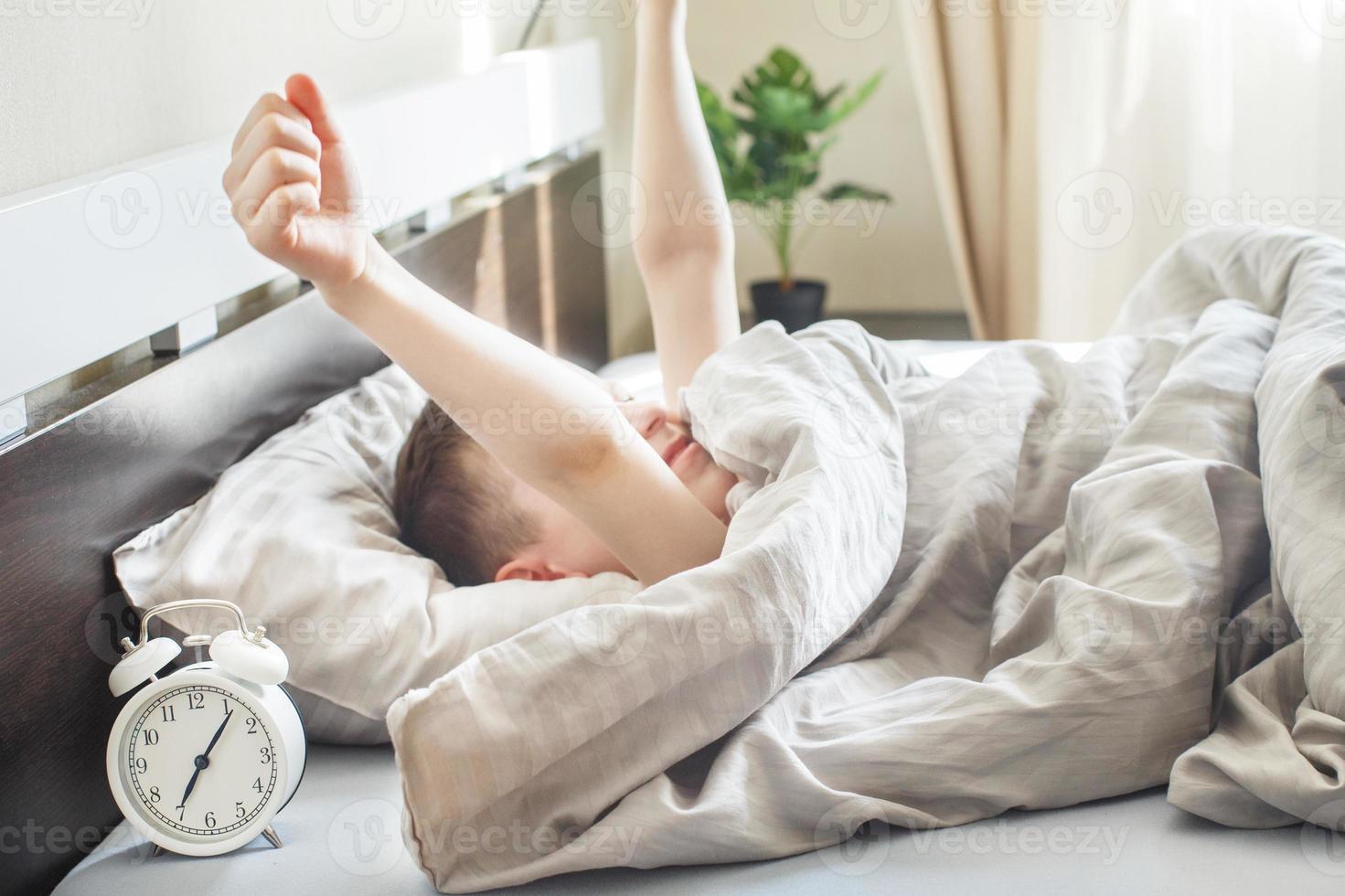 petit garçon allongé et s'étirant dans son lit à la maison le matin avec réveil photo