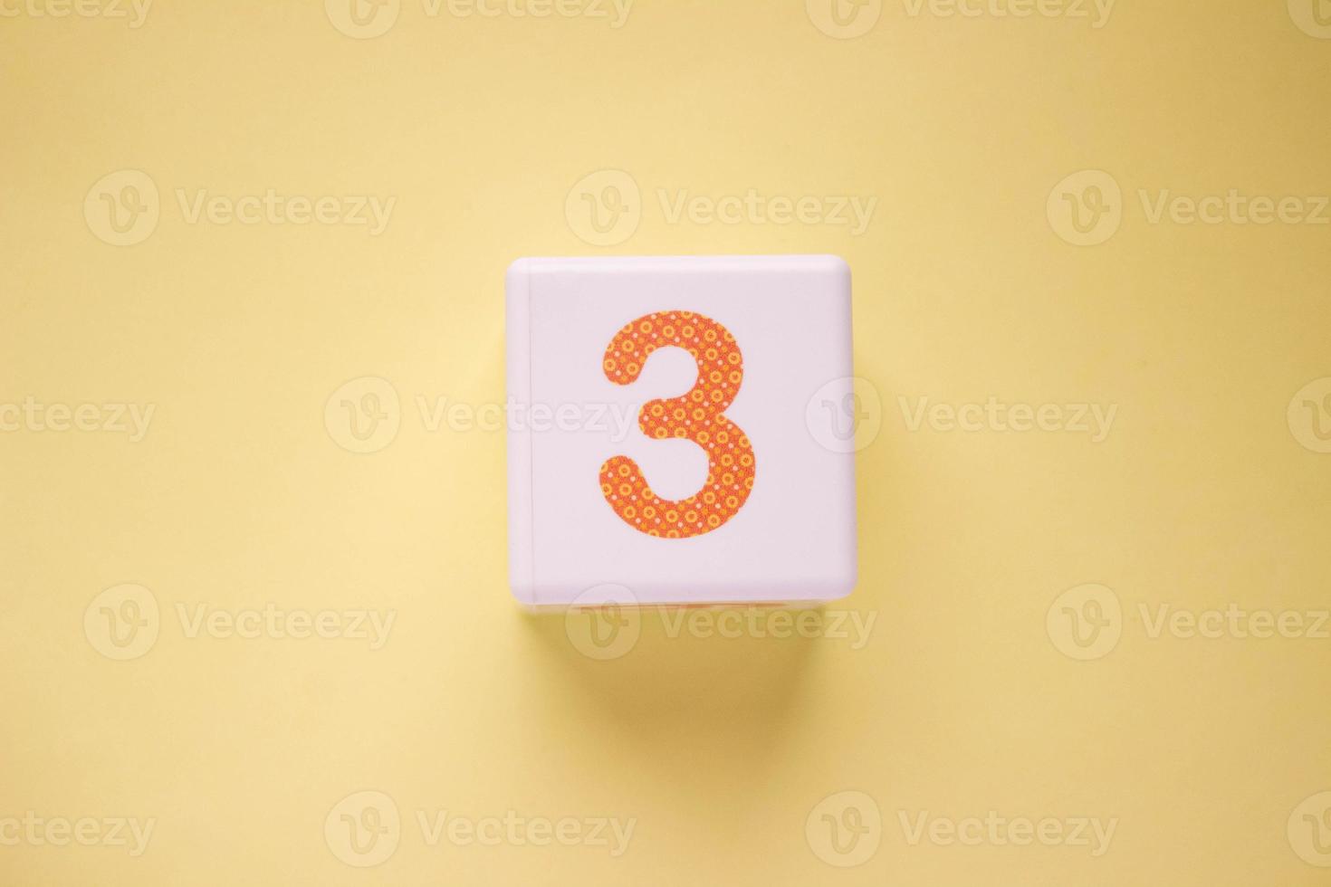 photo en gros plan d'un cube en plastique blanc avec un numéro orange 3 sur fond jaune. objet au centre de la photo