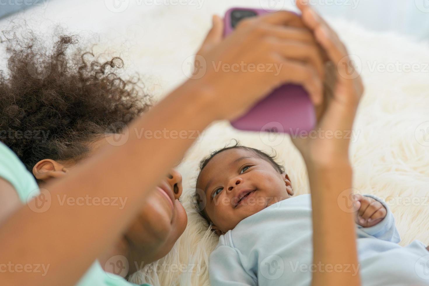 jeune mère afro-américaine et nouveau-né faisant un appel vidéo et souriant saluer. bonne sœur aînée prenant selfie avec petit frère bébé allongé sur le lit en regardant un téléphone portable souriant sans souci. photo
