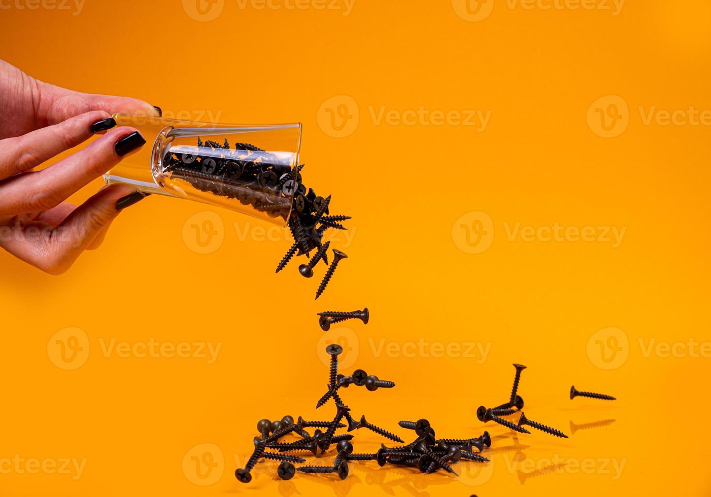 a laissé tomber de nombreuses vis autotaraudeuses métalliques en acier dans un verre, dans un pot seau. vis autotaraudeuse pour métal, pour fer, vis autotaraudeuse chromée, sur fond jaune orangé, photo