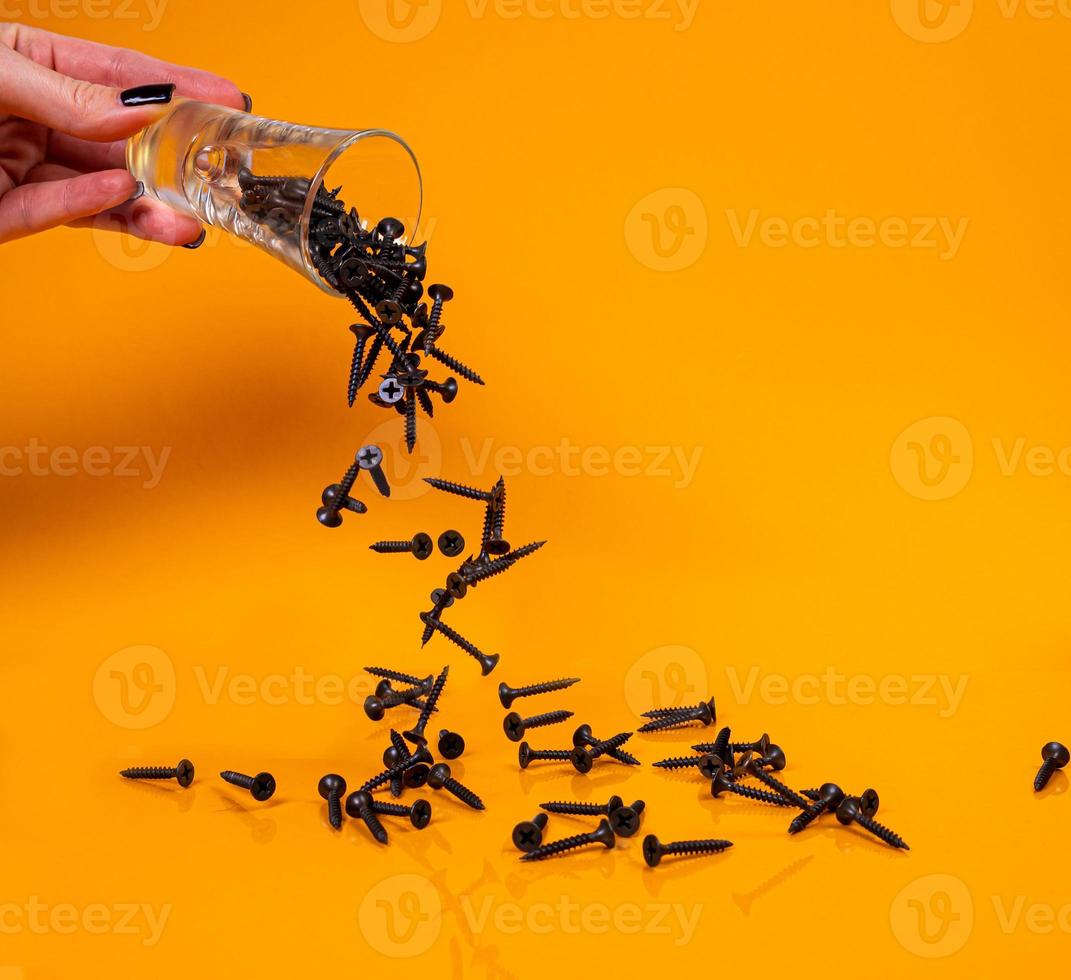 a laissé tomber de nombreuses vis autotaraudeuses métalliques en acier dans un verre, dans un pot seau. vis autotaraudeuse pour métal, pour fer, vis autotaraudeuse chromée, sur fond jaune orangé, photo