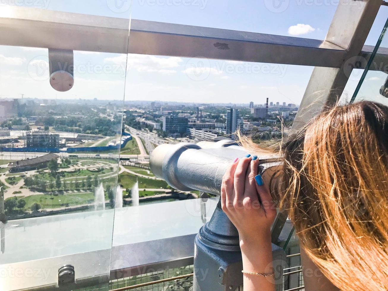 une belle fille regarde le paysage, un panorama de la ville dans la salle d'observation, une paire de jumelles, un télescope avec un accepteur de billets sur une terrasse d'observation en plein air photo