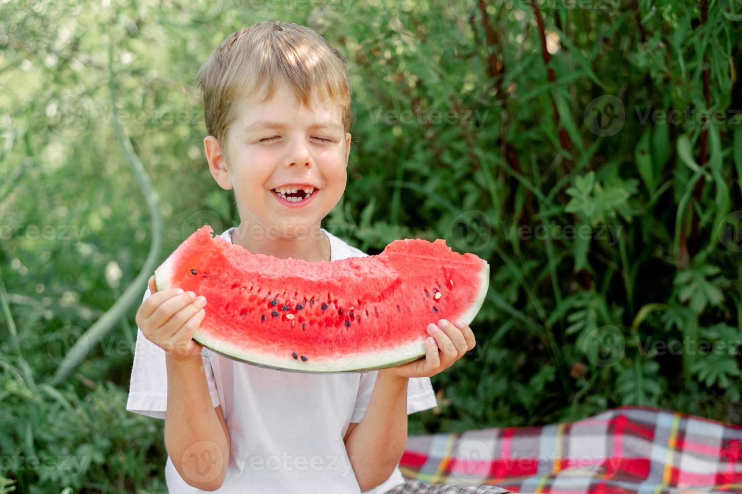 petit homme édenté mange un t-shirt blanc pastèque. pique-nique avec des pastèques. ambiance d'été. le garçon tient un gros morceau de pastèque dans ses mains. photo