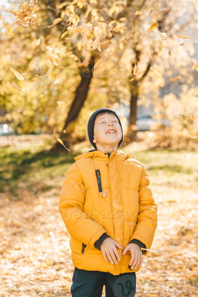 garçon dans une veste jaune, disperse les feuilles dans un parc en automne. l'enfant se réjouit des feuilles d'automne. enfance heureuse. veste et feuilles jaune vif photo