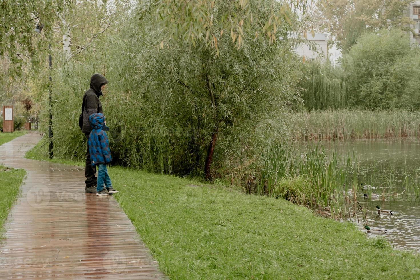 un homme avec un enfant dans une veste à capuche, se tenir debout et regarder le lac un jour de pluie photo