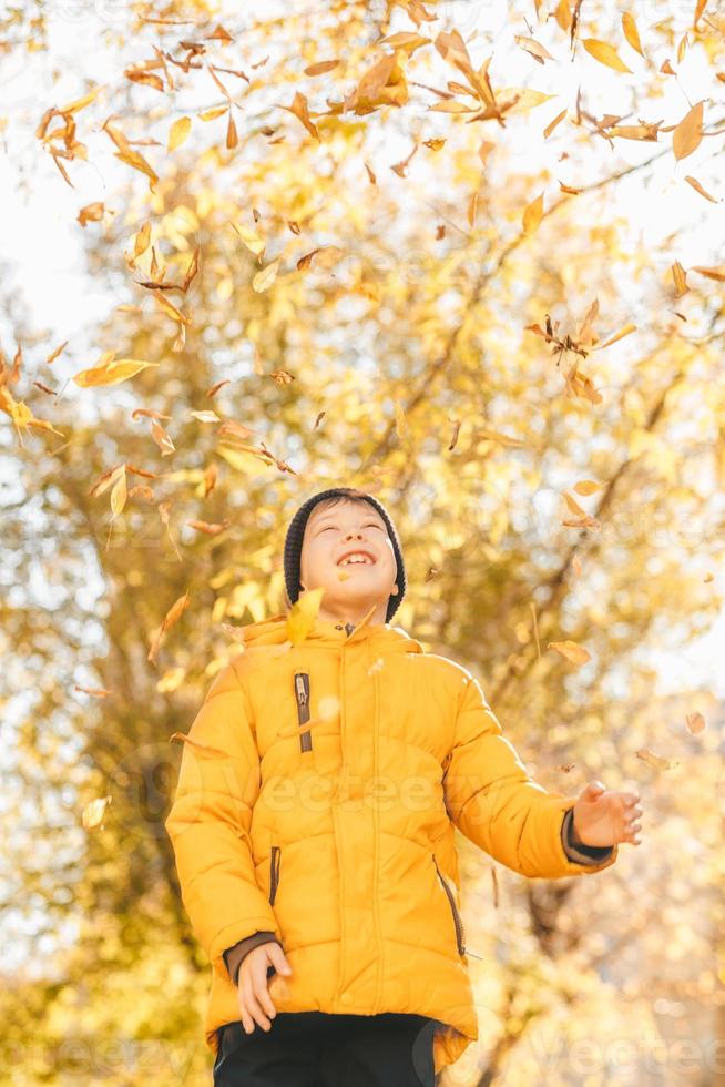 garçon dans une veste jaune, disperse les feuilles dans un parc en automne. l'enfant se réjouit des feuilles d'automne. enfance heureuse. veste et feuilles jaune vif photo