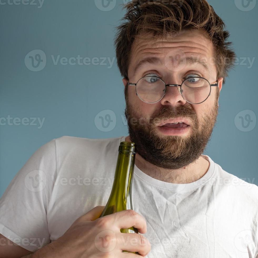 un homme mal rasé aux cheveux ébouriffés souffre d'une gueule de bois, il serre une bouteille de vin vide, fait une étrange grimace et a l'air négligé. photo