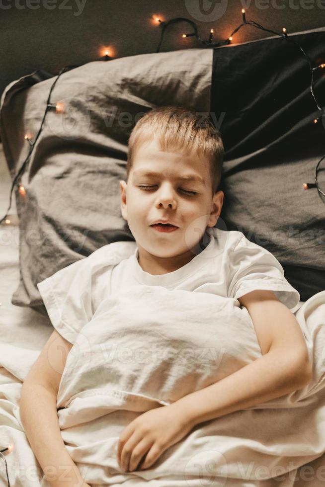 portrait d'un garçon dormant sous une couverture blanche et avec une guirlande. bébé de nuit de noël. petit garçon dormant sur un oreiller gris, cadre vertical photo