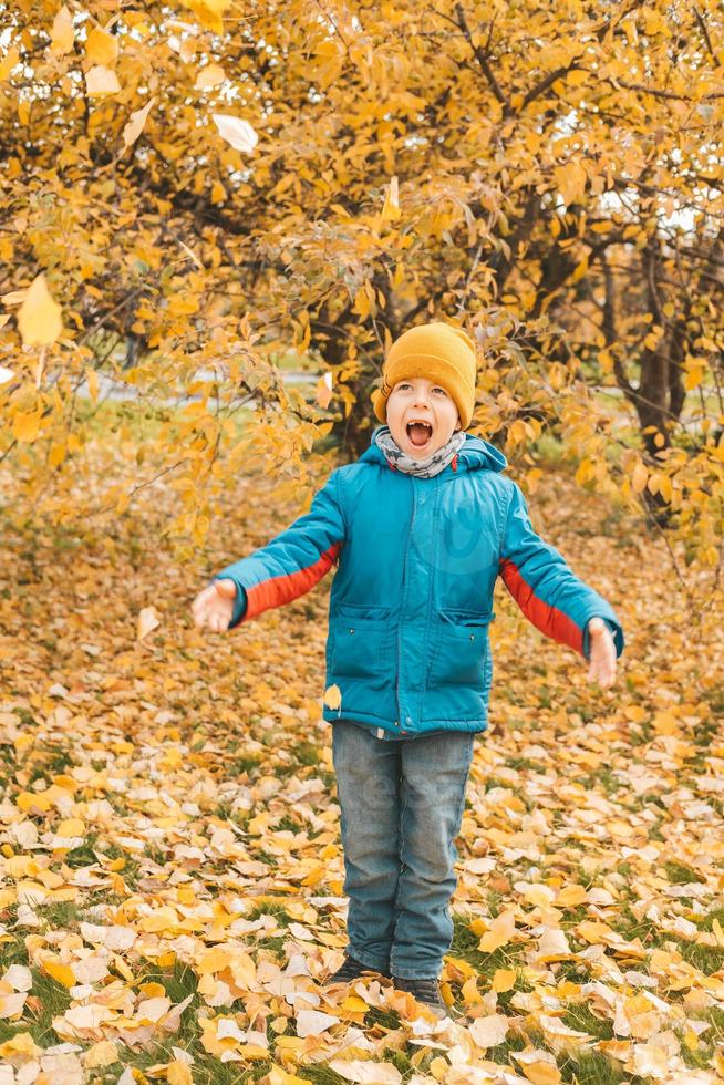 garçon dans une veste bleue, disperse les feuilles dans un parc en automne. l'enfant se réjouit des feuilles d'automne. enfance heureuse. veste et feuilles jaune vif. promenade avec les enfants dans le parc d'automne. photo