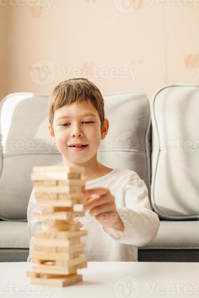 un garçon caucasien souriant joue à la maison, sur la table à jenga. jeux de société pour enfants et adultes. un passe-temps sans gadgets. jeux pendant les vacances à la maison. un enfant heureux joue avec des blocs de bois. photo