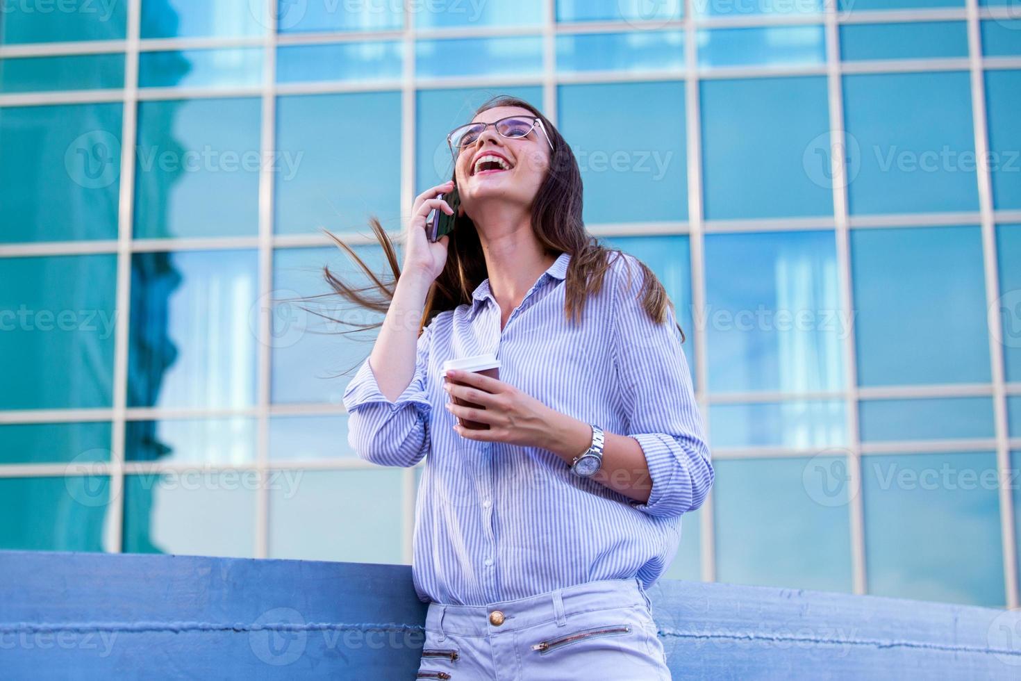 femme d'affaires souriante et heureuse parlant sur un smartphone mobile et buvant du café dans une tasse en papier jetable dans la rue avec des immeubles de bureaux en arrière-plan photo