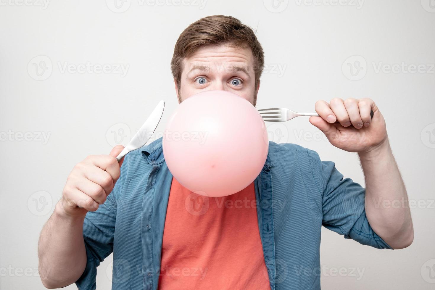 homme drôle tenant dans sa bouche un ballon et va l'éclater avec un couteau et une fourchette. fond blanc. photo