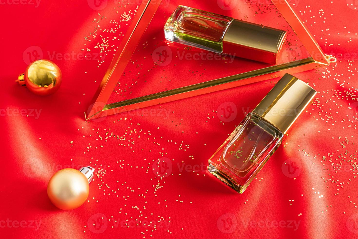 une bouteille de parfum transparente repose sur un tissu de satin rouge et se reflète dans un miroir avec des boules de noël. le concept d'un cadeau pour les vacances. photo