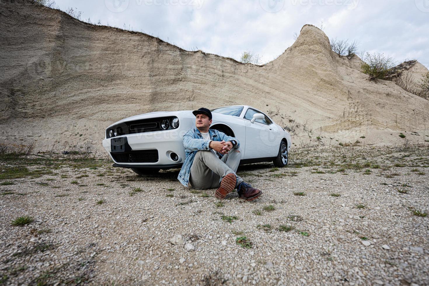 bel homme en veste de jeans et casquette est assis près de sa voiture de muscle blanche en carrière. photo