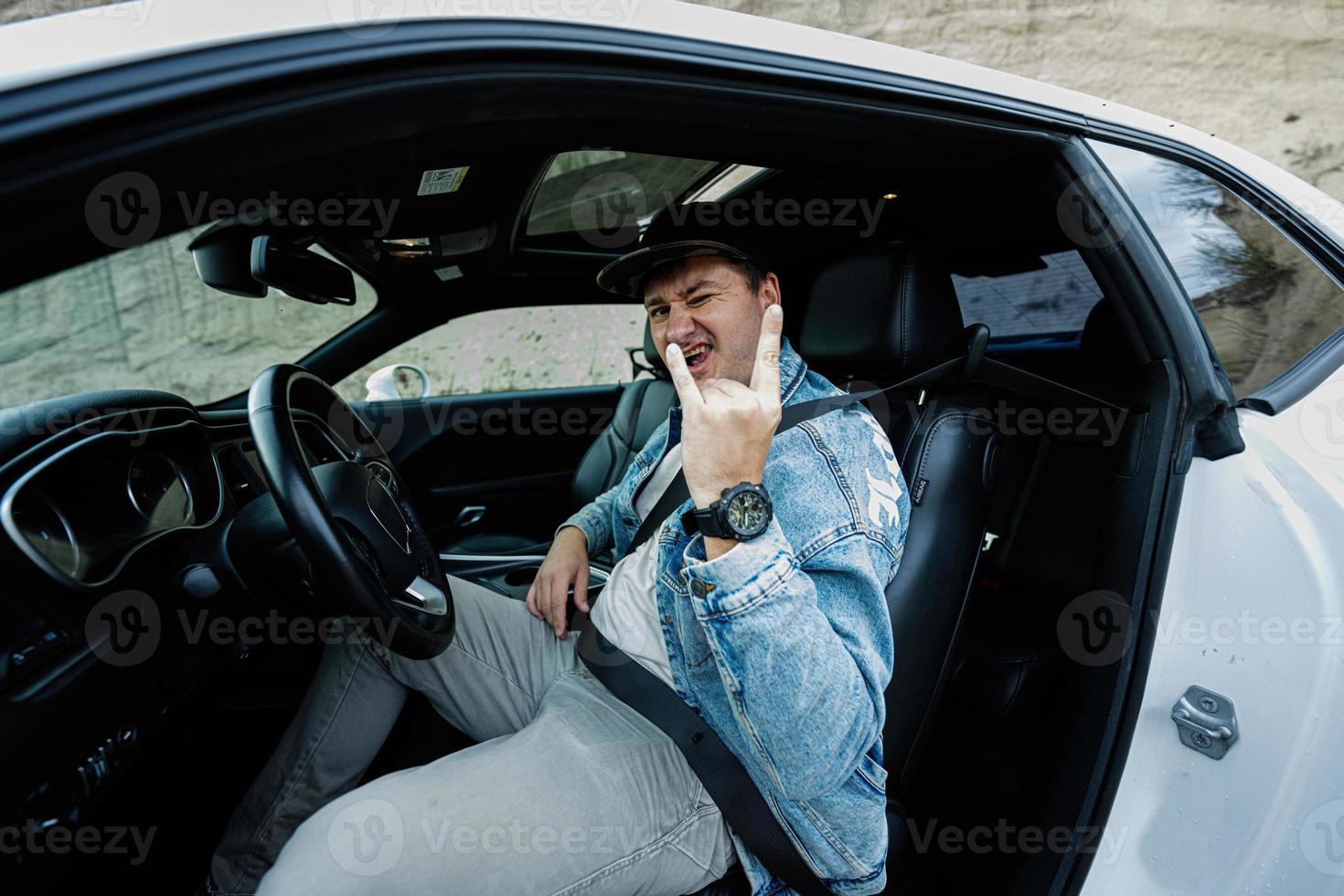bel homme en veste de jeans et casquette s'asseoir à sa voiture de muscle blanche et montrer le signe de la main rock and roll. photo