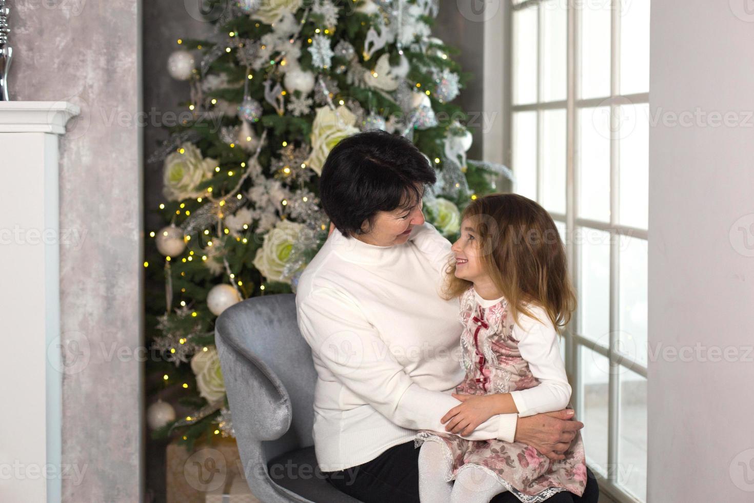 grand-mère avec une petite fille sur fond de décorations de noël et une grande fenêtre. vacances en famille, émotions, coffret cadeau. petite-fille sur les genoux de grand-mère. nouvel An photo