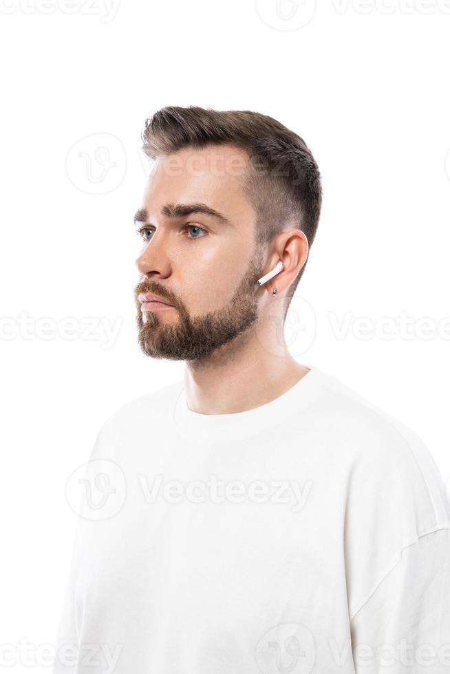 Bel homme barbu en vêtements blancs à l'aide d'écouteurs sans fil sur fond blanc photo