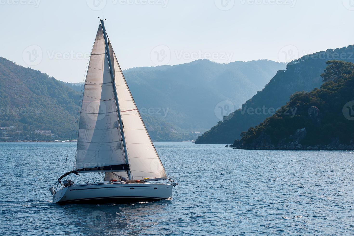 yacht à voile avec des voiles blanches sur une baie de mer ondulée sur fond de montagnes. photo