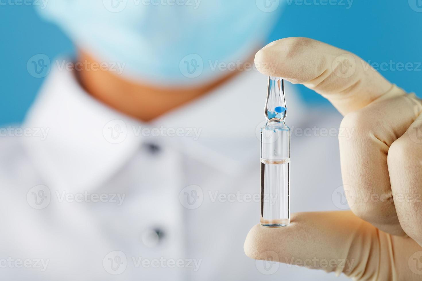 ampoule avec le vaccin contre le virus des maladies sur fond bleu entre les mains d'un médecin, scientifique en gants et masque. photo